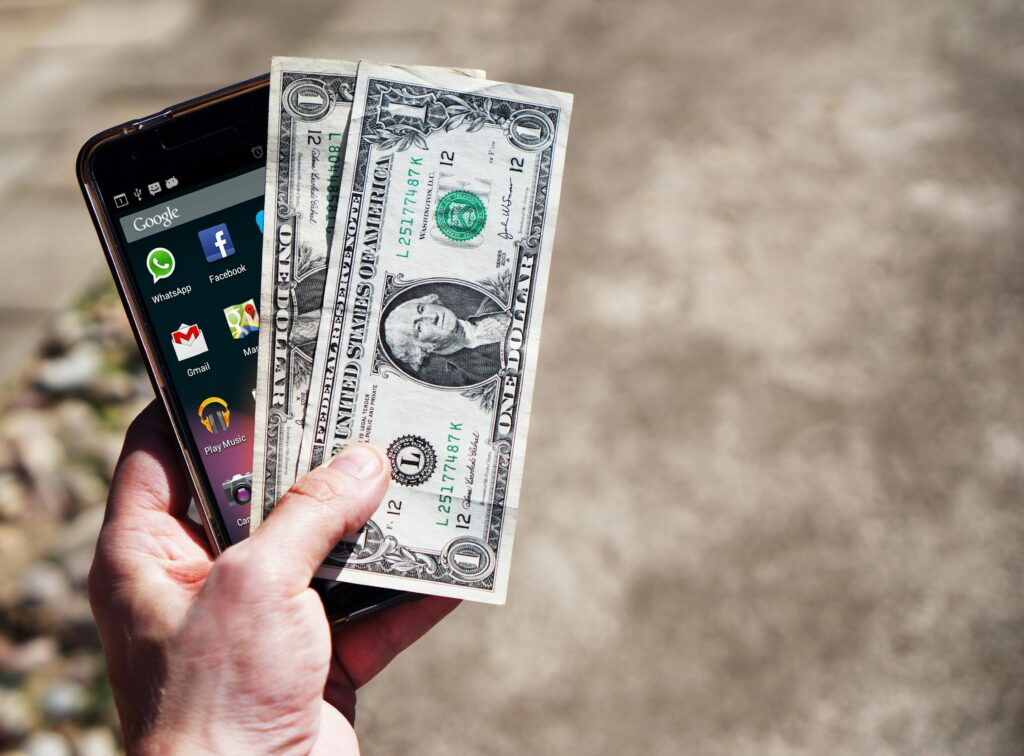 Close-up of a hand holding US dollar bills and a smartphone outdoors, showcasing financial technology, Facebook